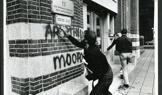 Zwart-wit foto waarop twee drie mannen te zien zijn voor de Zuid-Afrikaanse ambassade. Eén man schrijft met een spuitbus 'Apartheid is moord' op de muur. 